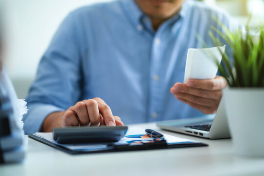 A man is sitting at a desk with a calculator and a receipt in his hand. He is focused on the receipt, possibly checking his expenses or trying to figure out how much he spent