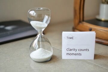 A clear hourglass with white sand flowing on a neutral tabletop next to a card about time