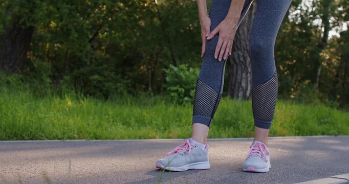 Woman runner suffering from kneecap pain. Crop adult sportswoman girl in running sportswear walking slowly on asphalt path road in green summer park stops to rub knee with hands. Sport injury concept