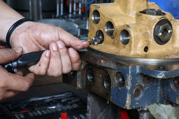Close-up of a worker using a screwdriver to remove a small screw from a heavy-duty hydraulic or industrial valve body during machine maintenance or overhaul.