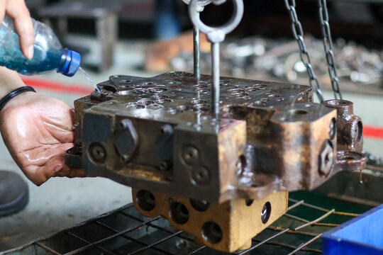 Worker rinsing a large, heavy hydraulic valve block with fluid from a bottle, a critical step during the cleaning process in an industrial equipment repair workshop.