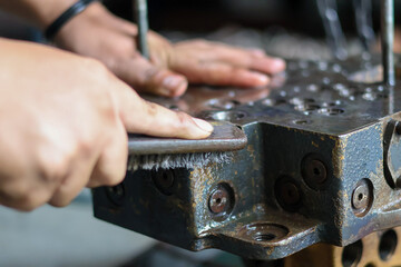 Close-up of a worker cleaning corrosion and grime from a heavy hydraulic valve block surface using a metal wire brush during equipment maintenance.