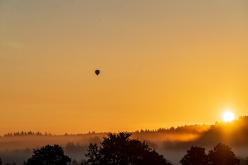 A hot air balloon floats gently as the sun sets, casting golden hues over the misty hills and trees
