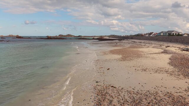Low drone footage of shoreline at the stunning Cobo Bay Guernsey Channel Islands with deserted sandy beach and lapping waves on a bright day with interesting sky