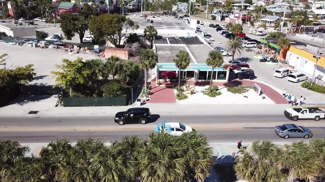 Aerial drone video of a tropical town with palm trees and cars driving as the camera lifts and rotates right to reveal the sandy beach and ocean waves and people swimming along the coast in Florida