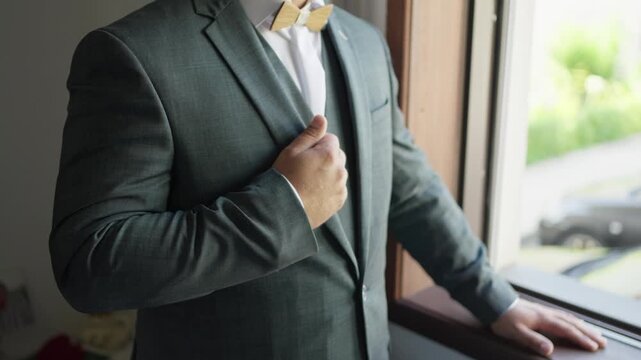 close up of groom fixing suit and bow tie near window before wedding