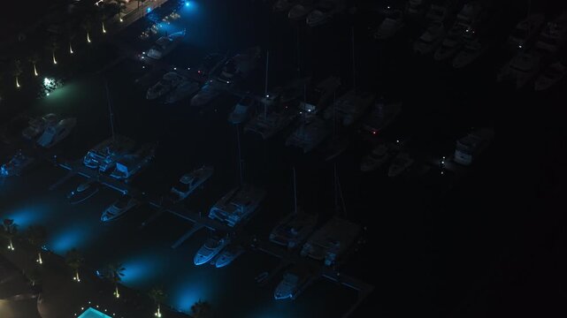 Aerial perspective from a tripod of luxury yachts and boats docked in a dark marina. Blue underwater lights illuminating tranquil harbor water at night