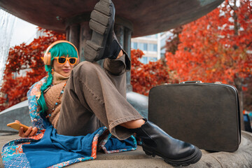 Young woman relaxes by a fountain with colorful headphones and vibrant hair, enjoying a calm moment during autumn