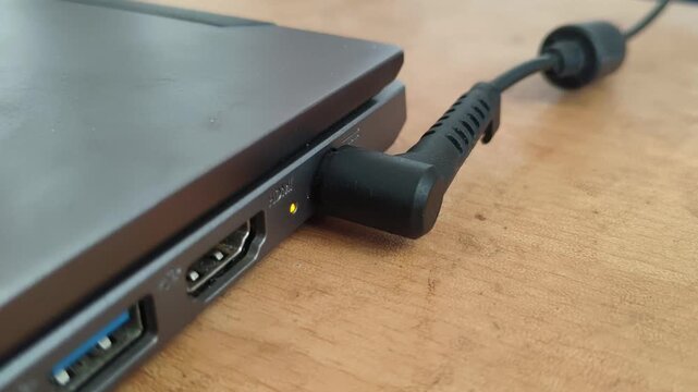 Hand plugging or connecting a old traditional round type black charger power cable cord into a laptop device charging port hole with glowing led light. Electricity or energy concept. Close up top view