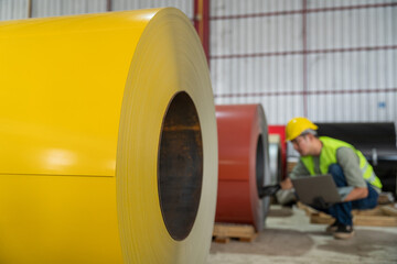 Close-up on a bright yellow metal coil roll in the foreground, with an industrial worker checking quality and inventory in the blurred background of the factory.