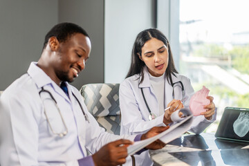 Two cardiologists engage in a detailed discussion on heart disease treatment, using an anatomical model to illustrate their points while consulting with a medical intern in a clinic, share knowledge