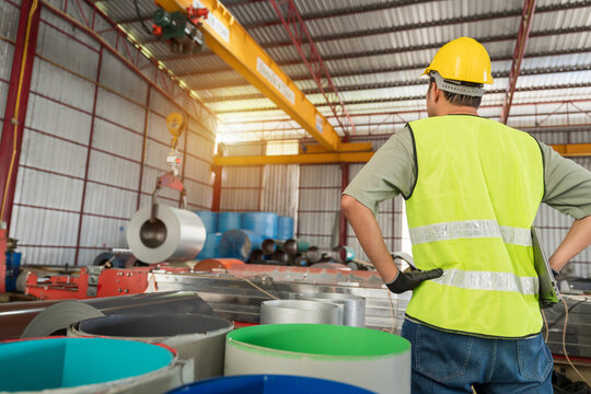 Industrial worker in safety uniform overseeing the operation of an overhead crane lifting a metal coil roll inside a bright factory warehouse.