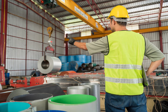 A Safety worker giving instructions and directing the movement of a large metal coil roll being lifted by an overhead crane in a factory.