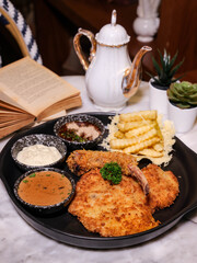 A delicious meal featuring a fried meat dish on a black plate with various sauces and fries, beautifully arranged. There is also a teapot and a book on the table