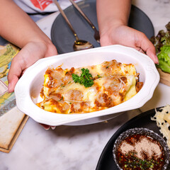 A delicious lasagna in a white serving dish is held by a pair of hands