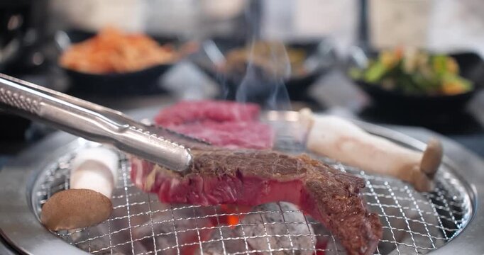 Close-up slow motion shot of metal tongs carefully flipping a strip of high-quality Hanu beef steak on a hot wire rack over glowing charcoal in a Korean restaurant