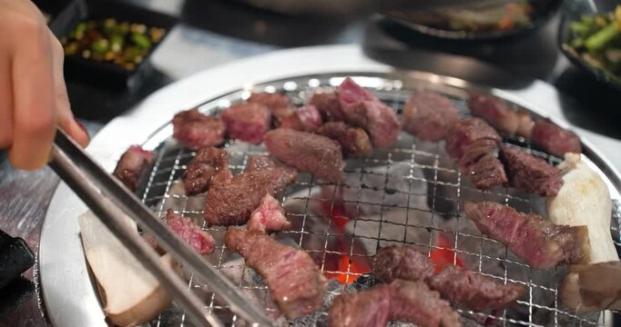 Close-up shot of tongs flipping multiple small, sizzling pieces of Hanu Korean beef and king oyster mushroom on a wire rack over hot glowing charcoal
