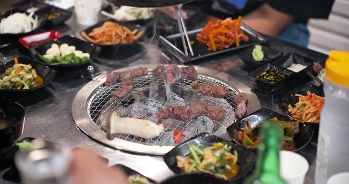 Wide-angle shot of a Korean restaurant tabletop filled with small bowls of colorful side dishes surrounding a central charcoal grill cooking hanu beef and king oyster mushrooms
