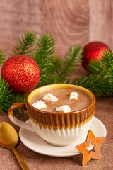 Cozy hot chocolate drink or sweet cocoa garnished with marshmallows served in cup on plate on dark brown table with red shiny christmas decoration baubles, wooden star decor and golden spoon for party