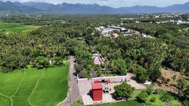 A serene aerial view captures a South Indian temple complex nestled within a landscape of lush greenery. A paved road winds through vast bright green paddy fields and coconut palm groves,