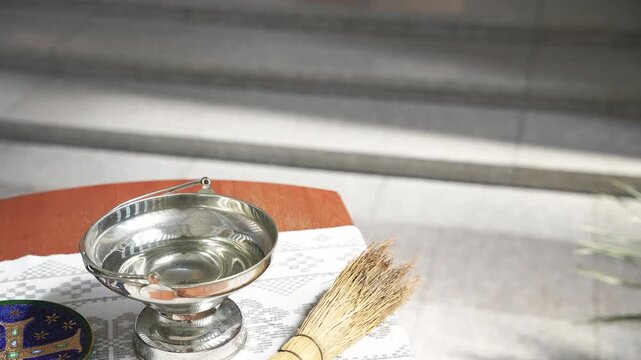 Holy water set with a silver vessel, brush, and decorative plate placed on a white cloth-covered wooden table inside a church, ready for use in a religious ceremony or blessing.