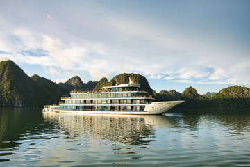 Large luxurious cruise ship sailing calm waters surrounded by towering limestone karsts and lush islands under a partly cloudy sky, offering scenic views and a tranquil travel experience