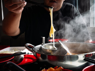 Person stretching melted cheese with chopsticks from steaming hot pot shabu divided metal pot on dining table