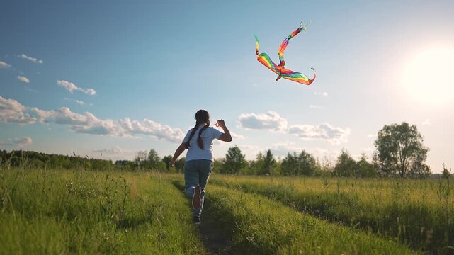 Girl running across green field outdoors. Kite with rainbow colors flying above grass. Summer play brings girl freedom. Girl holds kite high. Rainbow kite dances field. Happy girl runs in summer grass