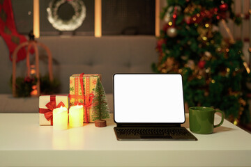 Blank display screen digital tablet and cup of hot chocolate on white table in decorated living room for Christmas