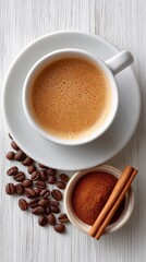 Fototapeta premium Overhead view of a white ceramic coffee cup filled with frothy coffee topped with cinnamon next to scattered dark roasted coffee beans and a cinnamon stick on a rustic white wooden surface.