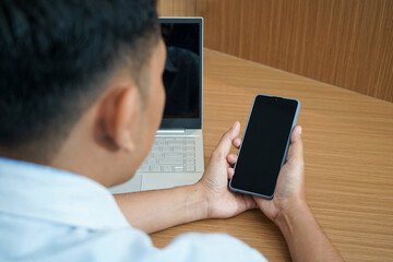 Close up of a male hands using a laptop and holding a smartphone with a blank screen, perfect for mockup or template design. Office desk background
