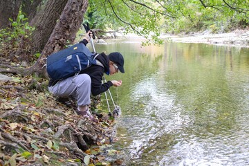  young female tourist with a backpack alone in nature drinks water from a river