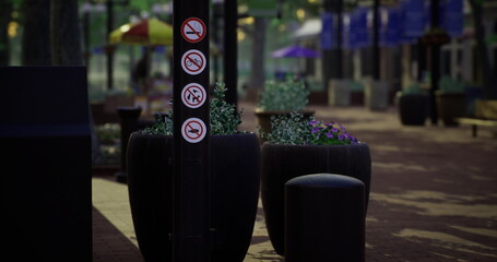 In a lively public plaza, multiple signs indicate restrictions on smoking, drinking, and pets. The area features planters filled with flowers and shaded seating under umbrellas.