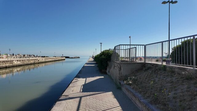 Long perspective view of a pedestrian walkway lining a calm port or canal entrance channel, leading toward the open sea. Features modern street lamps, safety railings, and the Italian flag under a bri