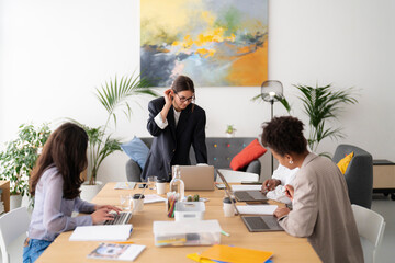 Three diverse women collaborate in a modern office setting. One stands, adjusting her glasses while reviewing a laptop. Two colleagues work on computers and documents at the table.