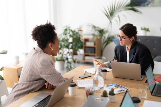 Two professional women collaborate during an office meeting. They sit at a wooden desk with laptops and documents, actively discussing business strategies in a bright workspace.