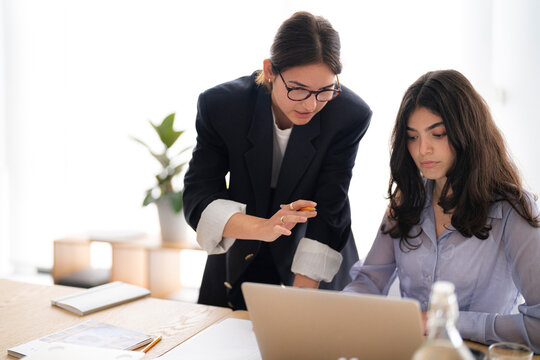 Two young businesswomen collaborate on a laptop in a modern office. One stands, pointing at the screen, while the other sits, focused on the task. They are engaged in a productive discussion.