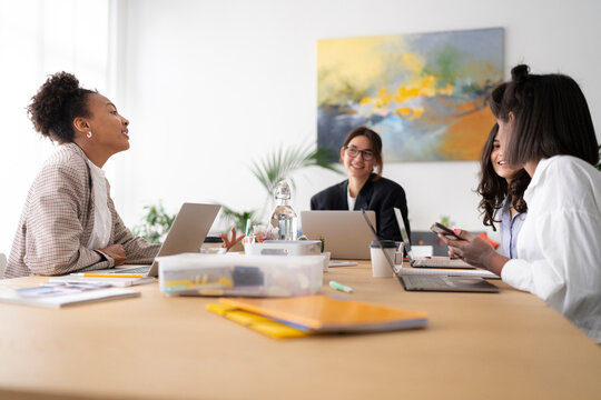 A diverse group of professional women collaborates in a bright office meeting. They are engaged in discussion, working on laptops, and sharing ideas around a large wooden table.