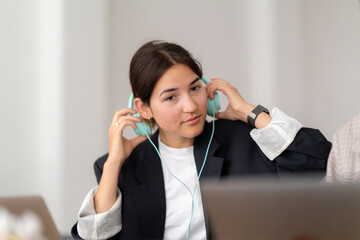 A young woman adjusts her light blue wired headphones, looking directly at the camera. She is dressed in a blazer and white shirt, engaged in an indoor activity.