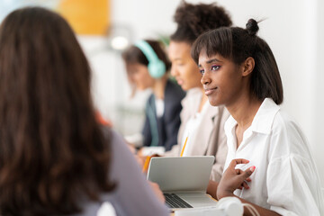 Young diverse women are engaged in a collaborative work or study session. One woman in a white shirt listens attentively while others use laptops and headphones in the background.