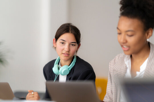 Diverse young women collaborate in a modern office. One focuses on her laptop with headphones around her neck, while the other discusses a project.