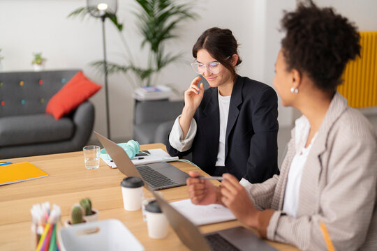 Two diverse businesswomen collaborate on laptops at a modern office desk. They are engaged in a productive discussion, sharing ideas and working efficiently.