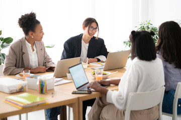 Four diverse women collaborate at a bright office table. They use laptops, discuss ideas, and enjoy a casual working lunch together. This scene shows teamwork and productivity.