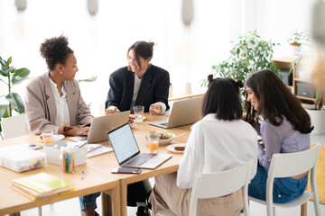 Four diverse young women are collaborating at a bright office table. They work on laptops, share ideas, and engage in cheerful discussion during a productive meeting.