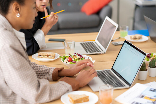 Two diverse women work on laptops at a shared desk, enjoying a healthy lunch of salads and sandwiches. They are focused on their tasks in a modern office setting.