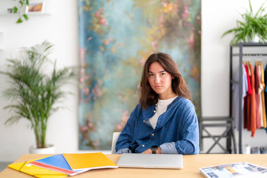 A young woman with a neutral expression sits at a wooden desk, a closed laptop and colorful folders in front of her. She looks directly at the viewer in a bright workspace.