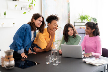 Four diverse young women gather around a laptop, collaborating and sharing ideas. They are smiling and engaged, working together at a table in a bright, modern setting.
