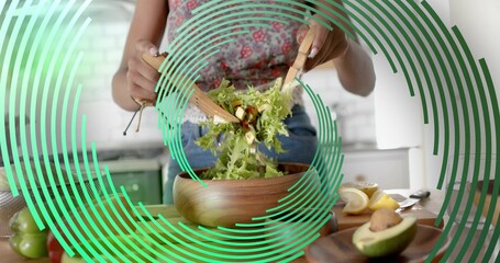 Home cook in floral top and jeans tossing salad at kitchen countertop, with wooden tongs, bowl