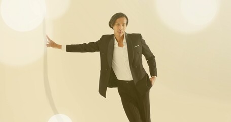 Leaning Asian model wearing black blazer and trousers extending left arm in studio, bokeh lights
