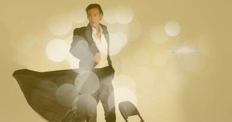 Standing Asian man wearing black suit and white shirt in studio, with folding chair, bokeh lights